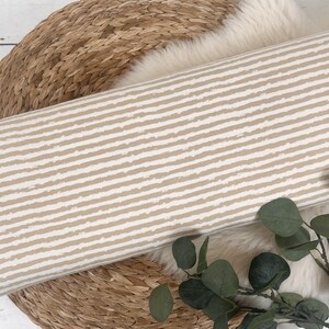 Pu&ograve; includere: Un tessuto a righe beige e bianco con un motivo testurizzato a pennellate. Il tessuto &egrave; arrotolato e posizionato su un cesto intrecciato.