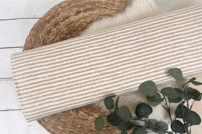 Pu&ograve; includere: Un tessuto a righe beige e bianco con un motivo testurizzato a pennellate. Il tessuto &egrave; arrotolato e posizionato su un cesto intrecciato.