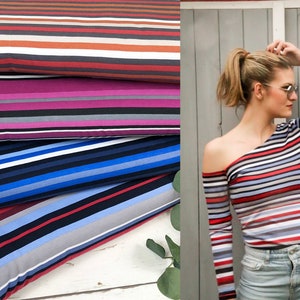 Può includere: Quattro rotoli di tessuto a righe in diverse combinazioni di colori. Il tessuto è piegato e impilato su una superficie di legno bianco. Il tessuto presenta una varietà di colori, tra cui rosso, blu, nero, bianco e grigio.