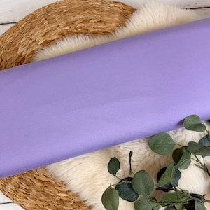 May include: A roll of solid lavender fabric. The fabric is smooth and appears to be a medium weight. The roll is resting on a white and brown surface with green foliage in the foreground. This fabric is suitable for sewing projects.