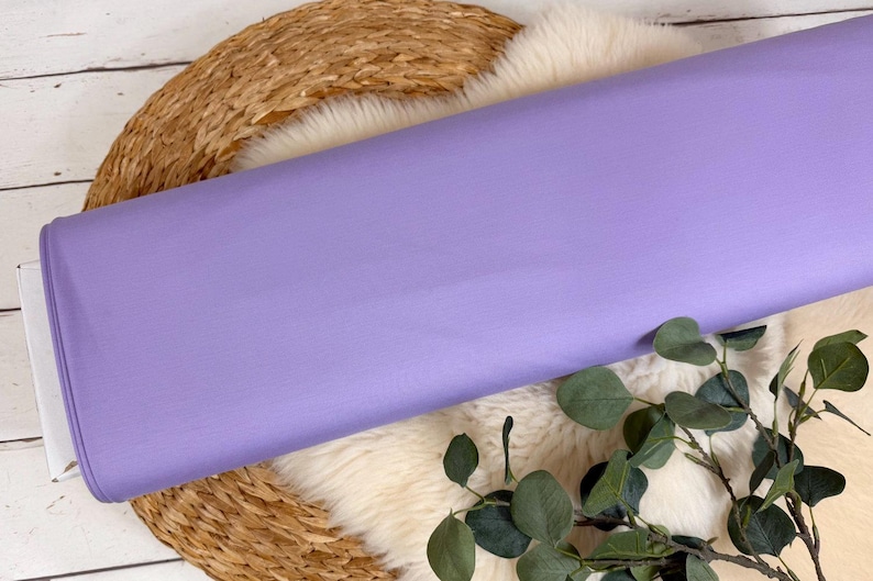 May include: A roll of solid lavender fabric. The fabric is smooth and appears to be a medium weight. The roll is resting on a white and brown surface with green foliage in the foreground. This fabric is suitable for sewing projects.