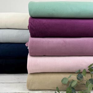 Op de afbeelding: Stapel van zes stofstalen in verschillende kleuren, waaronder beige, roze, lavendel, donkerblauw, lichtgrijs en groen. De stalen zijn gevouwen en verticaal gestapeld.
