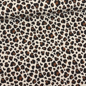 Può includere: Tessuto color crema con un motivo leopardato ripetuto. Il motivo presenta macchie marroni scure e marroni medie di varie dimensioni e forme. Il tessuto è piegato, mostrando la consistenza e il drappeggio del materiale. Adatto per abbigliamento e arredamento.