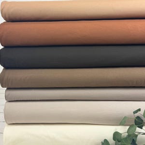 Op de afbeelding: Een stapel stoffen in verschillende neutrale kleuren, waaronder beige, bruin en zwart. De stof lijkt een zacht materiaal te zijn, mogelijk katoen of linnen. De afbeelding is goed belicht en toont de verschillende tinten en texturen van de stof.