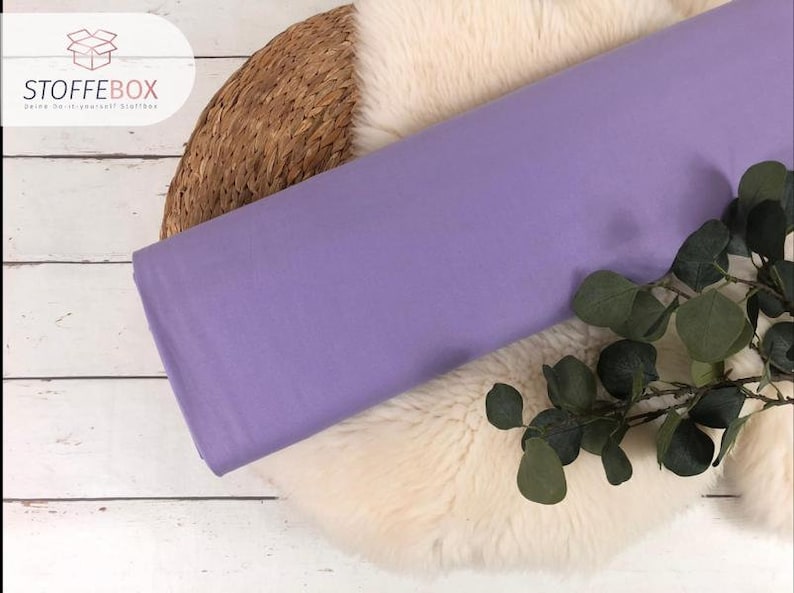 May include: A roll of light purple fabric on a white faux fur surface with a woven basket and green leaves.