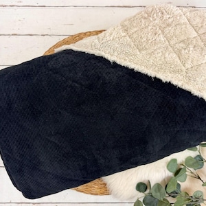 K&ouml;nnte beinhalten: Eine schwarze Corddecke mit cremefarbenem, gestepptem Sherpa-Futter. Die Decke ist gefaltet und zeigt beide Texturen. Der Hintergrund umfasst eine Holzoberfl&auml;che und einen Zweig mit Gr&uuml;n.
