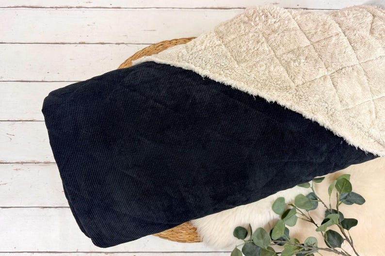 K&ouml;nnte beinhalten: Eine schwarze Corddecke mit cremefarbenem, gestepptem Sherpa-Futter. Die Decke ist gefaltet und zeigt beide Texturen. Der Hintergrund umfasst eine Holzoberfl&auml;che und einen Zweig mit Gr&uuml;n.