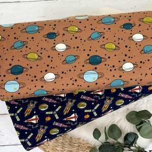 Op de afbeelding: Twee stofstalen met ruimtethema-patronen. De bovenste staal is bruin met witte, blauwe en gele planeten met ringen. De onderste staal is donkerblauw met witte, gele en oranje raketten en satellieten.