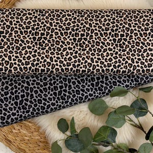 Puede incluir: Dos rollos de tela con estampado de leopardo. Uno es crema con manchas marrones, el otro gris con manchas negras. La tela se exhibe sobre una cesta tejida y una superficie blanca y esponjosa. Ideal para manualidades.