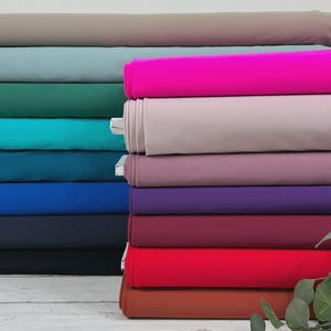 Peut inclure: Une pile de 14 rouleaux de tissu de différentes couleurs, dont turquoise, bleu, violet, rouge et marron. Le tissu est disposé en quinconce, le rouleau du haut étant légèrement décalé par rapport au rouleau du bas. Une plante verte feuillue se trouve au premier plan.