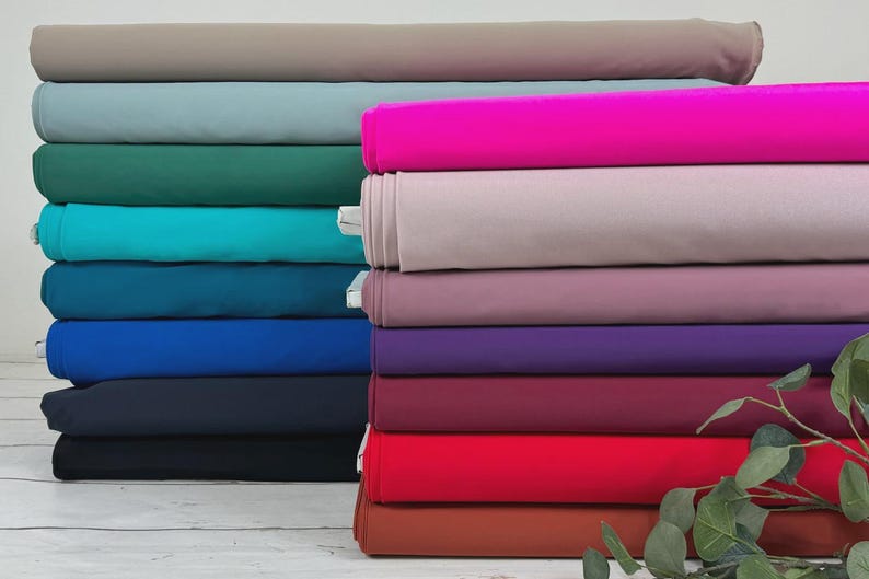 May include: A stack of 14 rolls of fabric in various colors, including teal, blue, purple, red, and brown. The fabric is arranged in a staggered pattern, with the top roll slightly offset from the bottom roll. A green leafy plant is in the foreground.