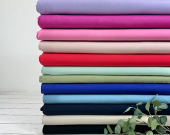 Könnte beinhalten: Ein vertikaler Stapel gefalteter Stoffballen in verschiedenen Unifarben, darunter Lavendel, Magenta, Pink, Creme, Rot, Minzgrün, Oliv, Königsblau, Hellblau, Marineblau, Beige und Schwarz, mit einem kleinen grünen Blattzweig an der Basis.