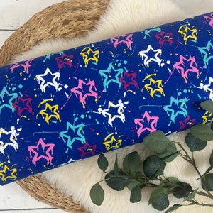 May include: Blue fabric with a white, pink, yellow, and turquoise star pattern. The stars are outlined in black and have paint splatters on them.