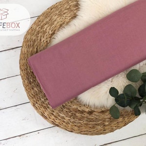 May include: A close-up of a dusty rose colored fabric swatch. The fabric is smooth and has a subtle sheen. The swatch is lying on a woven straw mat.