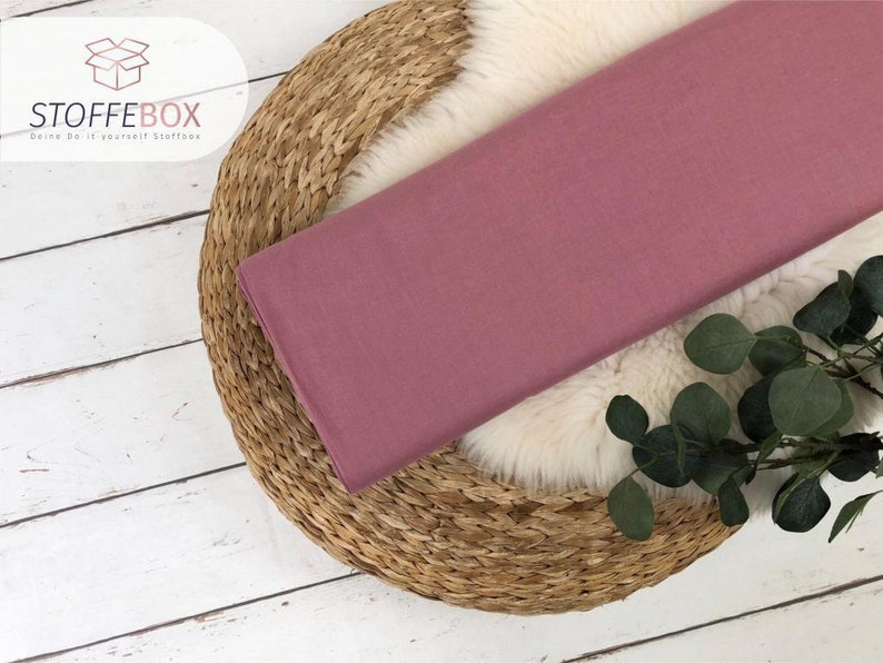 May include: A close-up of a dusty rose colored fabric swatch. The fabric is smooth and has a subtle sheen. The swatch is lying on a woven straw mat.