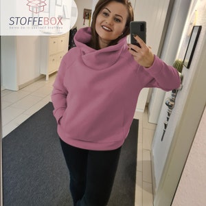 Puede incluir: Una sudadera rosa de gran tama&ntilde;o con cuello de pico y un bolsillo grande en la parte delantera. La sudadera la lleva puesta una persona en un reflejo de espejo.