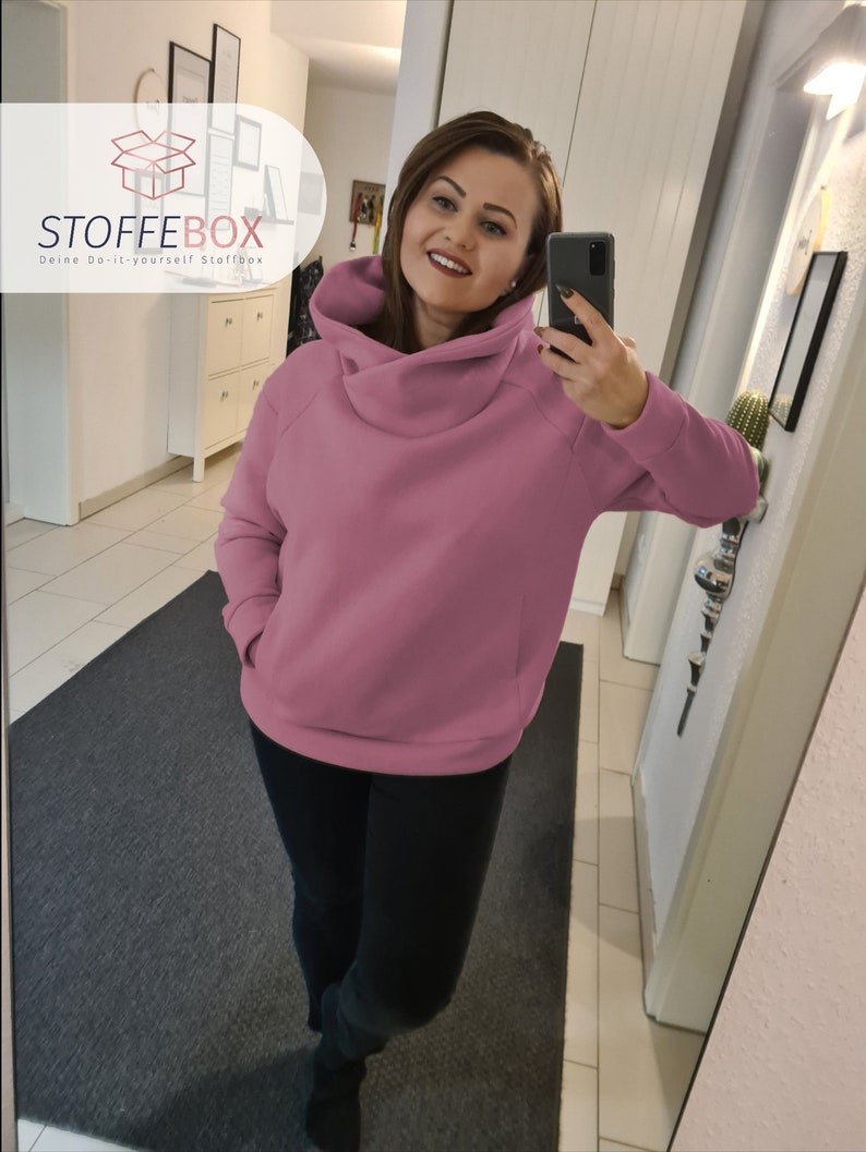Puede incluir: Una sudadera rosa de gran tama&ntilde;o con cuello de pico y un bolsillo grande en la parte delantera. La sudadera la lleva puesta una persona en un reflejo de espejo.
