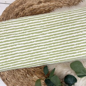 Pu&ograve; includere: Un tessuto a righe verde e bianco con un motivo testurizzato a pennellate. Il tessuto &egrave; appoggiato su una superficie intrecciata.