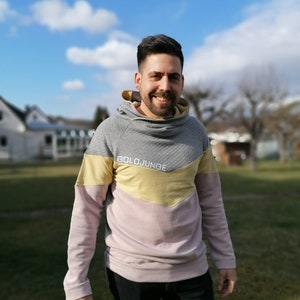 Puede incluir: Un hombre lleva una sudadera con capucha gris, rosa y amarilla con el texto "BOLDJUNGE" en la parte delantera. Est&aacute; de pie en una zona cubierta de hierba con un cielo azul y nubes blancas en el fondo.