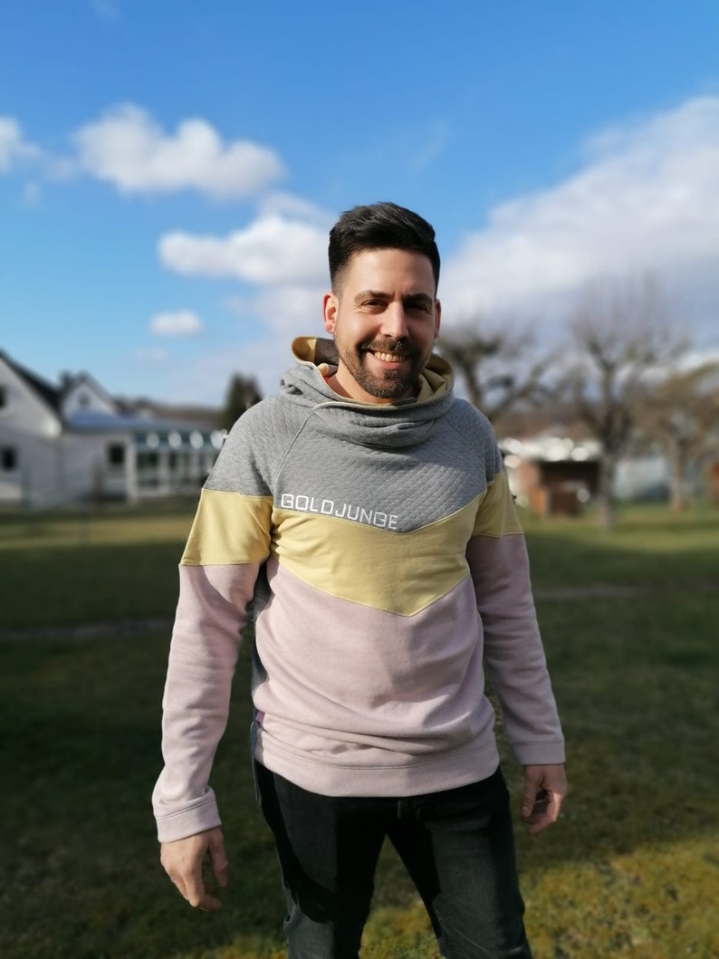 Puede incluir: Un hombre lleva una sudadera con capucha gris, rosa y amarilla con el texto "BOLDJUNGE" en la parte delantera. Est&aacute; de pie en una zona cubierta de hierba con un cielo azul y nubes blancas en el fondo.