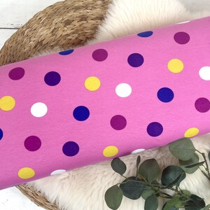 May include: Pink fabric with a polka dot pattern in yellow, white, purple, and blue. The fabric is rolled up and sitting on a woven basket.
