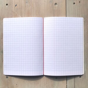 Puede incluir: Un cuaderno abierto con p&aacute;ginas cuadriculadas. Las p&aacute;ginas son blancas con un patr&oacute;n de cuadr&iacute;cula azul claro. La encuadernaci&oacute;n es roja y el cuaderno est&aacute; abierto sobre una superficie de madera clara.