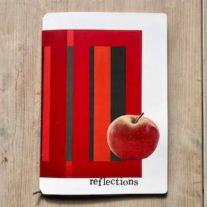 Puede incluir: Una manzana roja descansa sobre un bloc de notas rectangular blanco con un dise&ntilde;o abstracto rojo y negro. La palabra "reflections" est&aacute; impresa en negro en la parte inferior. El bloc de notas est&aacute; sobre una superficie de madera.