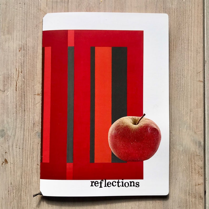 Puede incluir: Una manzana roja descansa sobre un bloc de notas rectangular blanco con un dise&ntilde;o abstracto rojo y negro. La palabra "reflections" est&aacute; impresa en negro en la parte inferior. El bloc de notas est&aacute; sobre una superficie de madera.