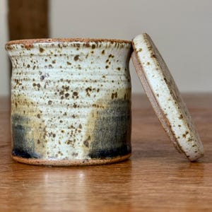 Può includere: Un barattolo cilindrico in ceramica con coperchio abbinato appoggiato di lato. L'esterno presenta uno smalto color crema e bianco maculato nella parte superiore, che sfuma in un tono blu-grigio scuro e spento alla base con striature visibili.