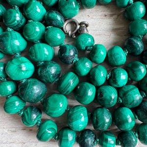 May include: A necklace composed of numerous polished, rounded malachite beads in varying shades of deep and vibrant green, featuring distinct natural banded patterns. The beads are strung together and secured with a silver-toned metal spring ring clasp.