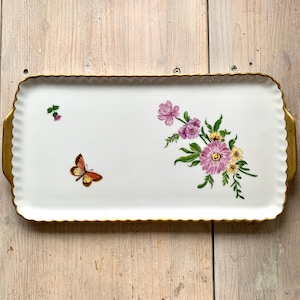 Könnte beinhalten: Ein rechteckiges Serviertablett in Weiß mit goldfarbenem Rand und einem floralen Design. Das Design zeigt rosa, gelbe und violette Blumen mit grünen Blättern und einem einzelnen orangefarbenen Schmetterling.