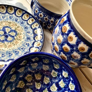 Può includere: Una collezione di stoviglie in ceramica blu e bianca. Il set comprende un piatto, una ciotola e due tazze. I disegni presentano un motivo ripetuto di punti marroni e bianchi e motivi floreali. I pezzi sono disposti su una superficie chiara.