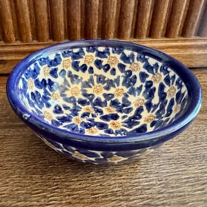 Può includere: Una piccola ciotola rotonda in ceramica con un motivo dipinto a mano di petali di fiori blu scuro con centri marrone chiaro su uno sfondo color crema. La ciotola presenta un bordo blu scuro solido e una finitura liscia e lucida all'interno.