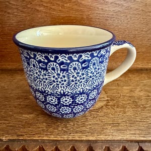 Puede incluir: Una taza de cerámica con un patrón floral azul y blanco. La taza tiene un interior color crema y un asa curvada. El exterior presenta un diseño detallado y simétrico en tonos azules y blancos.