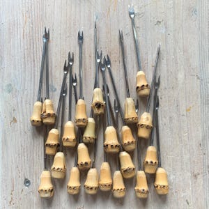 Könnte beinhalten: Eine Sammlung von Vintage-Cocktailgabeln mit Edelstahlzinken und hellbraunen Bambusgriffen. Die Gabeln sind auf einer hellen Holzoberfläche angeordnet, was ihr einzigartiges Design und ihre Handwerkskunst hervorhebt.