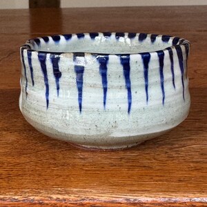 Può includere: Una ciotola in ceramica con una base grigio chiaro maculata e una serie di strisce verticali blu scuro a forma di goccia che si estendono dal bordo verso il basso. La ciotola presenta una forma arrotondata e leggermente irregolare.