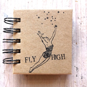 Könnte beinhalten: Ein kleines braunes Notizbuch mit einer schwarzen Spiralbindung. Der Umschlag zeigt eine Schwarzweißillustration einer springenden Frau mit erhobenen Armen, darunter der Text "FLY HIGH". Der Hintergrund des Umschlags ist mit kleinen schwarzen Sternen verziert.