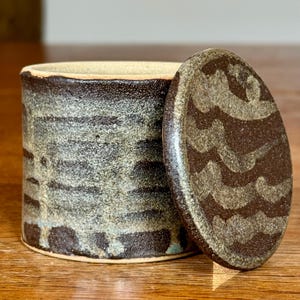 Op de afbeelding: Een cilindrische keramische pot met een getextureerde, donkerbruine en grijs gespikkelde afwerking, inclusief een bijpassend rond deksel. Het deksel heeft een golvend, lichtgekleurd patroon op een donkerbruine achtergrond en de pot heeft banden.