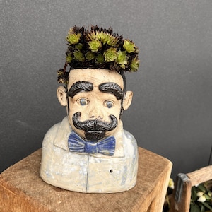 Könnte beinhalten: Ein Keramik-Blumentopf in Form eines Männerkopfes mit Schnurrbart und blauer Fliege. Der Topf ist mit grünen Sukkulenten gefüllt, was ein einzigartiges Dekorationsobjekt ergibt. Er steht auf einer Holzoberfläche.