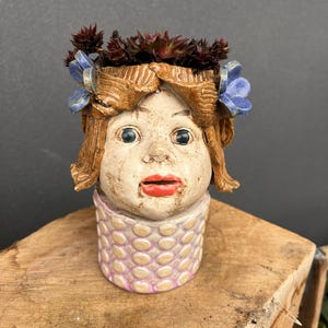 Könnte beinhalten: Keramik-Blumentopf in Form eines Mädchenkopfes mit braunen Haaren und blauen Blumenakzenten. Der Topf hat einen roten Mund und ist mit einer Sukkulentenpflanze gefüllt. Der Sockel ist hellrosa mit einem strukturierten Muster.