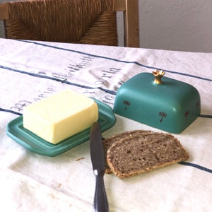 Könnte beinhalten: Eine tealfarbene Butterdose aus Keramik mit einer goldenen Krone auf dem Deckel und Palmenmotiven. Die Dose ist mit einer Butterstange gefüllt und ein Buttermesser liegt daneben auf dem Tisch. Zwei Scheiben dunkles Brot liegen auf dem Tisch.
