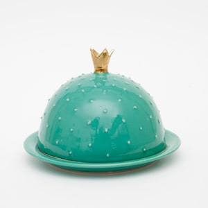 Könnte beinhalten: Türkisgrüne Keramik-Butterdose mit gewölbtem Deckel und goldener Krone. Der Deckel ist mit kleinen, weißen Punkten bedeckt. Die Dose steht auf einem passenden türkisfarbenen Teller. Einzigartiges Küchenutensil.