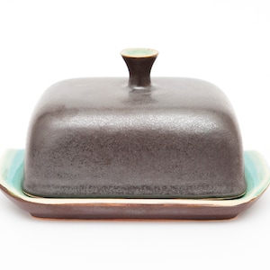 Könnte beinhalten: Ein Keramik-Butterdose mit einem dunkelgrauen, quadratischen Deckel und einem türkisfarbenen und braunen Boden. Der Deckel hat einen kleinen, abgerundeten Griff mit grünem Rand. Die Schale ist zum Servieren von Butter bestimmt.