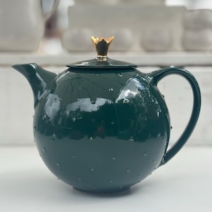 Könnte beinhalten: Eine dunkelgrüne Teekanne mit einem goldenen kronenförmigen Deckel. Die Teekanne hat einen runden Körper und einen kleinen Ausguss. Die Teekanne steht auf einer weißen Oberfläche.