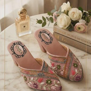 May include: A pair of dusty pink mules with a block heel, featuring intricate multi-colored floral embroidery and metallic thread detailing. The interior footbed is light pink and displays the brand name SNJLA in a decorative black oval frame.