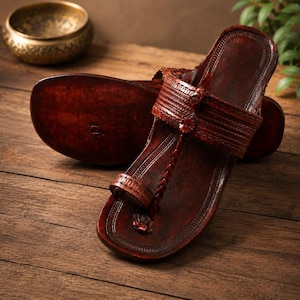 May include: A pair of dark brown leather sandals featuring a toe loop design, a wide strap across the midfoot with intricate embossed detailing, and a flat sole. The sandals have a polished finish and a structured shape with visible stitching.