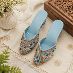 May include: A pair of light blue mules featuring a pointed toe and a block heel. The exterior is decorated with intricate multi-colored floral embroidery, sequins, and beadwork in shades of pink, yellow, green, and orange across the front.