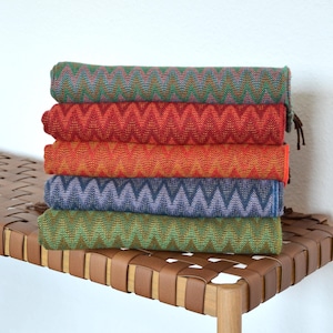 Könnte beinhalten: Ein Stapel aus fünf bunten Schals mit einem Chevron-Muster. Die Schals sind gefaltet und auf einer geflochtenen Lederbank gestapelt.