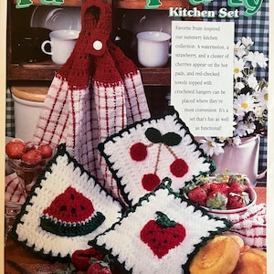 Op de afbeelding: Een keuken set met een leuk, fruitig thema. De set bevat een rood-witte geruite handdoek met een gehaakte hanger en drie witte gehaakte pannenlappen met watermeloen-, kersen- en aardbei-ontwerpen. De tekst "Fun 'N Fruity Kitchen Set" staat bovenaan.