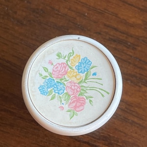 May include: A round, white compact mirror with a floral design. The design features pink, blue, and yellow flowers with green leaves on a cream background. The compact is set against a dark wood surface.